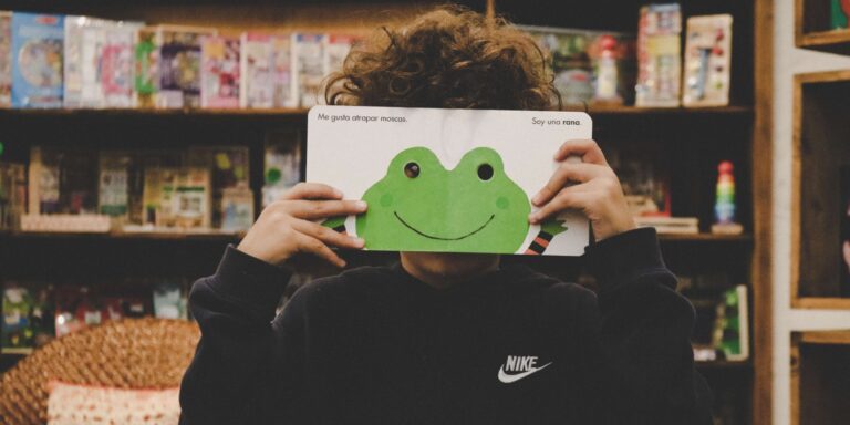 child holding up a book open to an illustration of a frog and holding it over their eyes